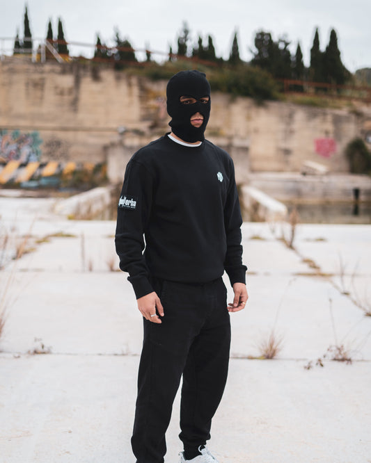 Modelo con un jersey negro de NonVerba, un pasamontañas y unos panatalones negro en una zona abandonada mirando a la cámara.